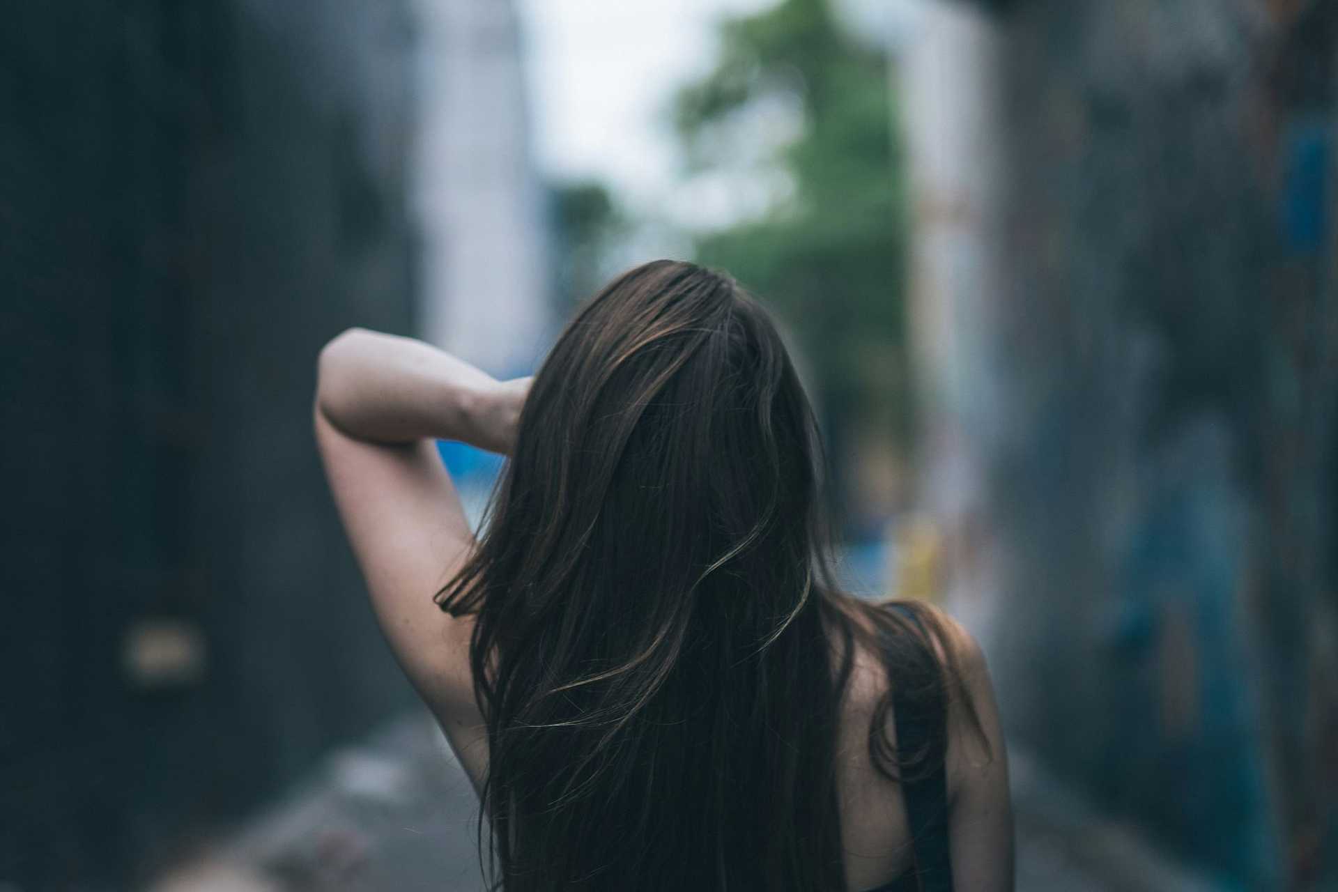 A woman with long hair standing in an alleyway, facing away.