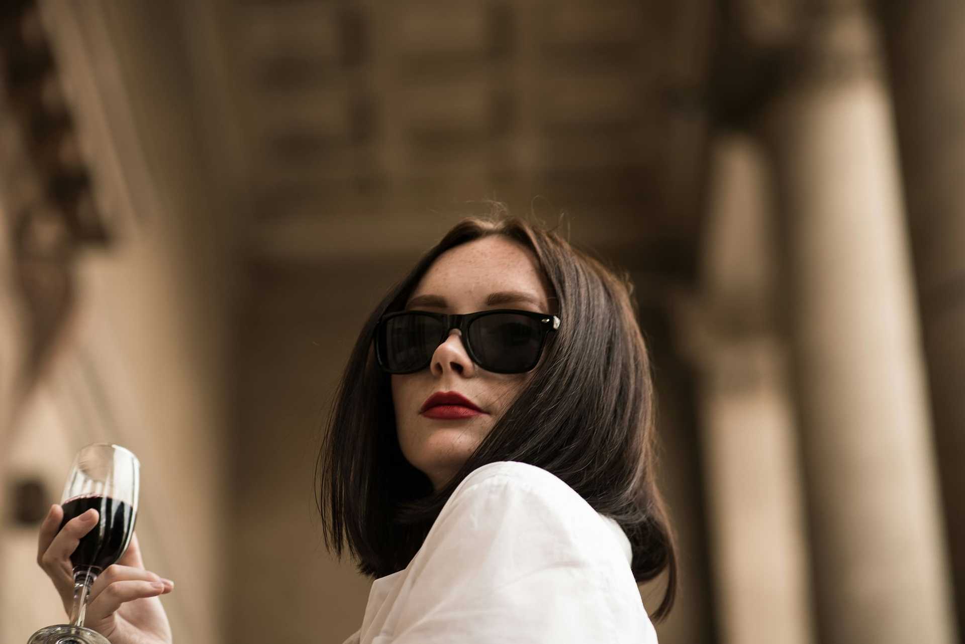 Woman in sunglasses holding a wine glass, standing in an elegant architectural setting.